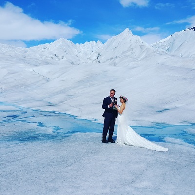 A bride and groom standing on a glacier with snow-covered mountains in the background.