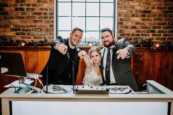 A DJ booth with turntables and a mixer, featuring a bride and groom posing with a DJ at a wedding venue.