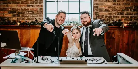 A DJ booth with turntables and a mixer, featuring a bride and groom posing with a DJ at a wedding venue.