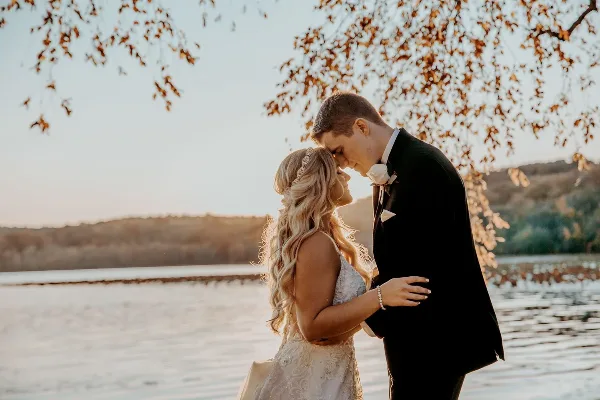 A bride and groom standing close together outdoors near a body of water, with the groom's forehead touching the bride's.