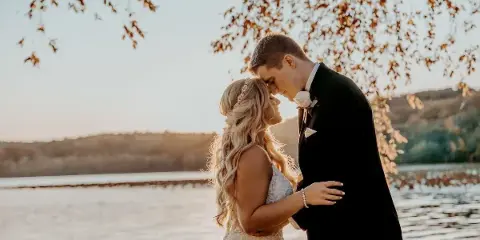 A bride and groom standing close together outdoors near a body of water, with the groom's forehead touching the bride's.