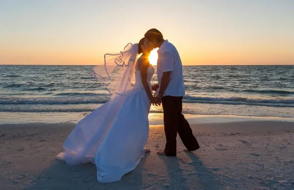 A bride and groom kissing on a beach at sunset.