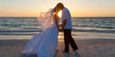 A bride and groom kissing on a beach at sunset.