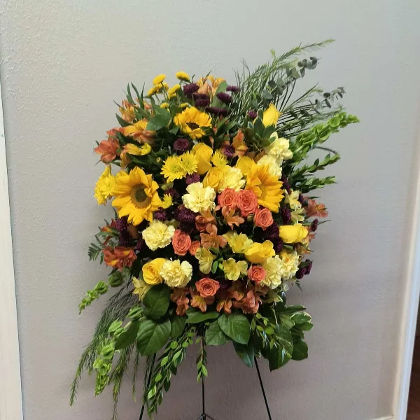A large, vibrant floral spray featuring yellow sunflowers, orange roses, and greenery arranged on a black easel stand.