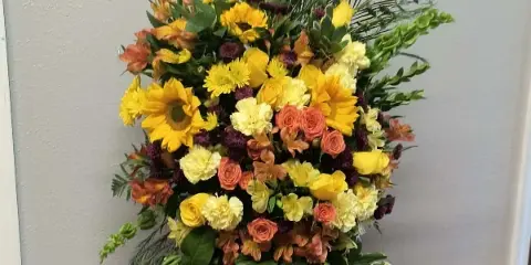 A large, vibrant floral spray featuring yellow sunflowers, orange roses, and greenery arranged on a black easel stand.