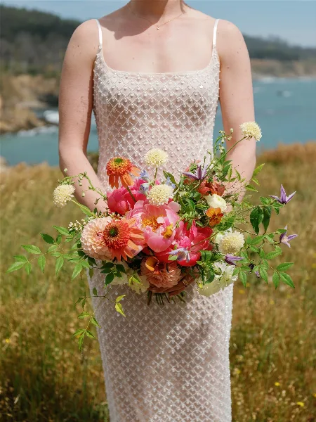 A bride in a white beaded dress holding a vibrant bouquet of pink peonies, orange dahlias, and purple clematis in a grassy field.
