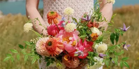 A bride in a white beaded dress holding a vibrant bouquet of pink peonies, orange dahlias, and purple clematis in a grassy field.