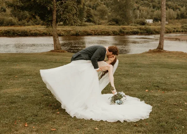 A bride and groom posing on a grassy lawn with a river and trees in the background.