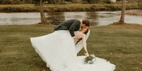 A bride and groom posing on a grassy lawn with a river and trees in the background.