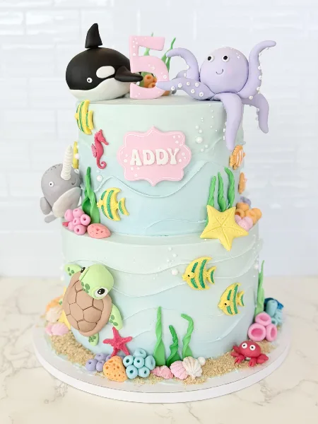 A two-tier light blue custom cake decorated with fondant sea creatures including a whale, octopus, and turtle, topped with a number 5 candle.