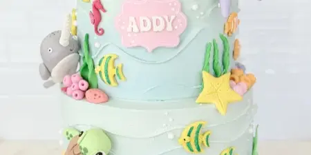 A two-tier light blue custom cake decorated with fondant sea creatures including a whale, octopus, and turtle, topped with a number 5 candle.