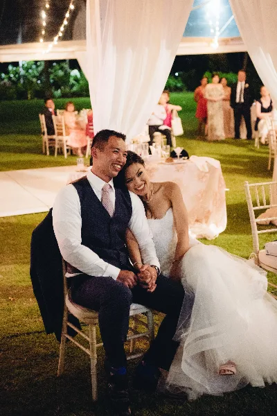 A bride and groom sitting together at an outdoor wedding reception under a draped canopy with string lights.