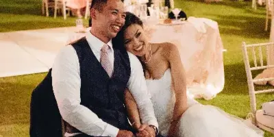 A bride and groom sitting together at an outdoor wedding reception under a draped canopy with string lights.