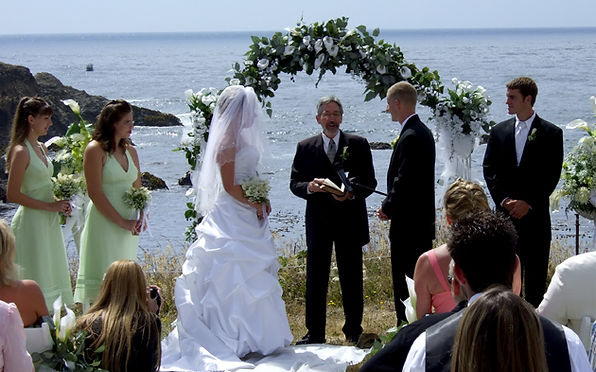 A wedding ceremony taking place outdoors overlooking the ocean, with the bride, groom, and officiant standing under a floral arch.