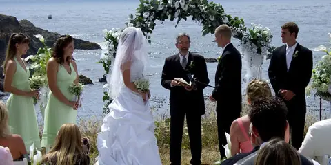 A wedding ceremony taking place outdoors overlooking the ocean, with the bride, groom, and officiant standing under a floral arch.