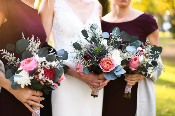 A bride and two bridesmaids in maroon dresses holding floral bouquets with pink roses and eucalyptus.