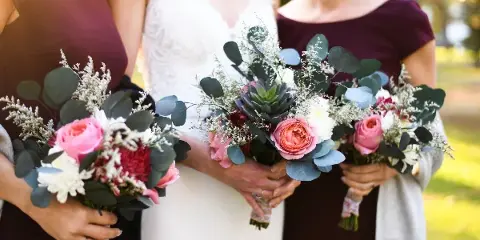 A bride and two bridesmaids in maroon dresses holding floral bouquets with pink roses and eucalyptus.