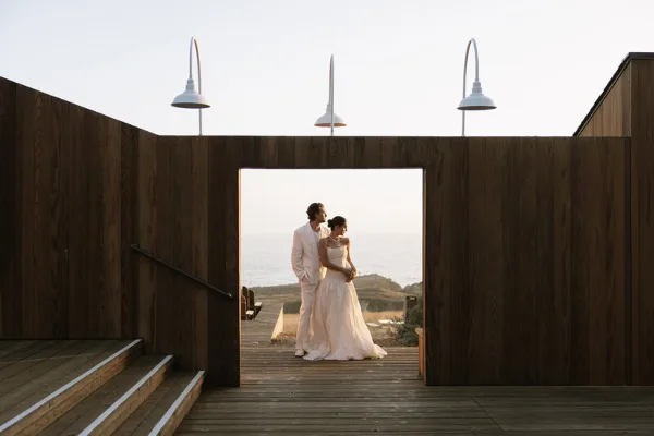A bride and groom standing together in a wooden architectural frame overlooking the ocean.