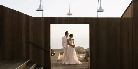 A bride and groom standing together in a wooden architectural frame overlooking the ocean.