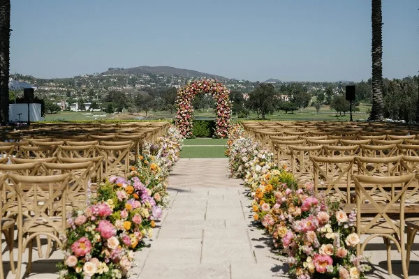 Outdoor wedding ceremony setup featuring a floral arch and aisle arrangements with rows of wooden chairs.
