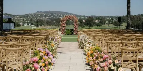 Outdoor wedding ceremony setup featuring a floral arch and aisle arrangements with rows of wooden chairs.