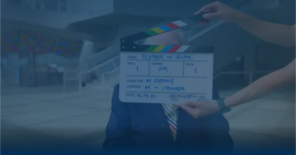 A person holding a film clapperboard in front of a man in a suit, representing a video production set.