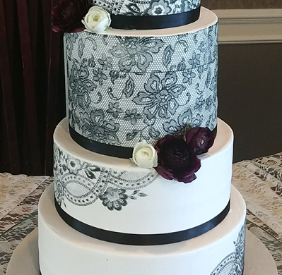 A three-tiered white wedding cake featuring black lace patterns, black ribbon bands, and dark red and white floral accents.