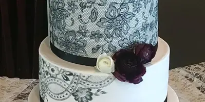 A three-tiered white wedding cake featuring black lace patterns, black ribbon bands, and dark red and white floral accents.