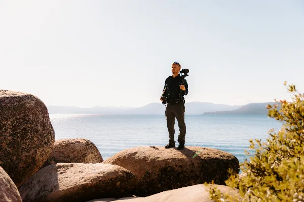 A videographer standing on large rocks by a body of water, holding a professional camera rig.