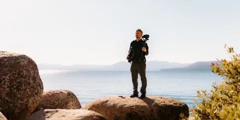 A videographer standing on large rocks by a body of water, holding a professional camera rig.
