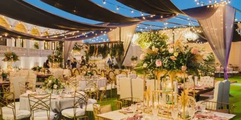 An outdoor evening banquet area with tables, gold chairs, and floral centerpieces under hanging fabric and string lights.