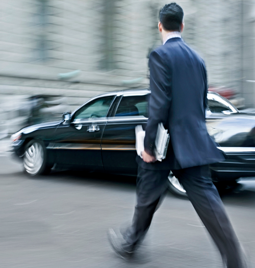 A man in a business suit walking toward a black limousine on a city street.
