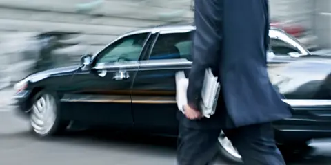 A man in a business suit walking toward a black limousine on a city street.