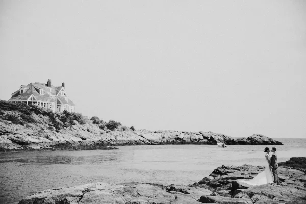 Black and white photo of a bride and groom standing on a rocky coast with a large house in the background.