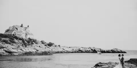 Black and white photo of a bride and groom standing on a rocky coast with a large house in the background.