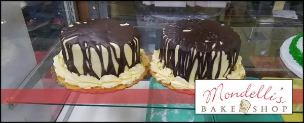 Two large chocolate and cream cakes displayed in a glass bakery case with the Mondelli's Bake Shop logo.