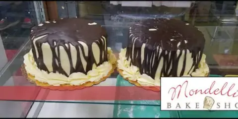 Two large chocolate and cream cakes displayed in a glass bakery case with the Mondelli's Bake Shop logo.
