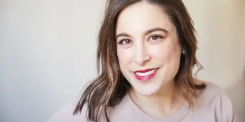 Portrait of a smiling woman with brown hair and red lipstick, wearing a light pink top.