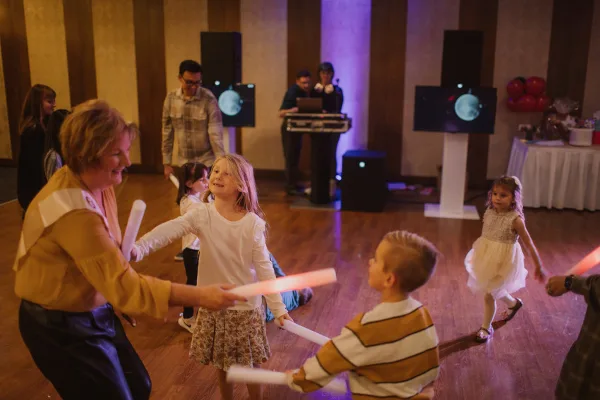 A DJ booth with professional speakers and a laptop is visible in the background while children and adults dance with glow sticks at an indoor event.