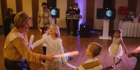 A DJ booth with professional speakers and a laptop is visible in the background while children and adults dance with glow sticks at an indoor event.