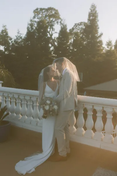 A bride and groom in wedding attire standing on a balcony with a white balustrade, sharing a kiss under a veil with a bouquet of flowers.