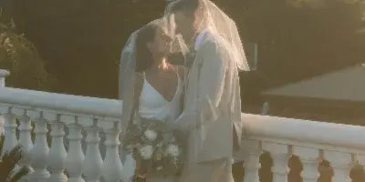 A bride and groom in wedding attire standing on a balcony with a white balustrade, sharing a kiss under a veil with a bouquet of flowers.