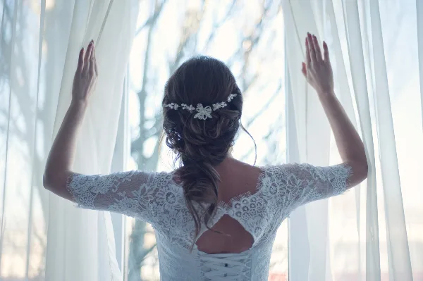 A bride in a white lace wedding dress with an updo hairstyle and floral hair accessory, standing with her back to the camera and hands raised to hold open sheer white curtains.