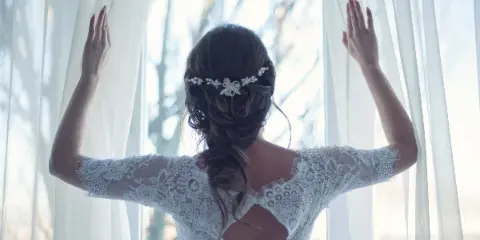 A bride in a white lace wedding dress with an updo hairstyle and floral hair accessory, standing with her back to the camera and hands raised to hold open sheer white curtains.