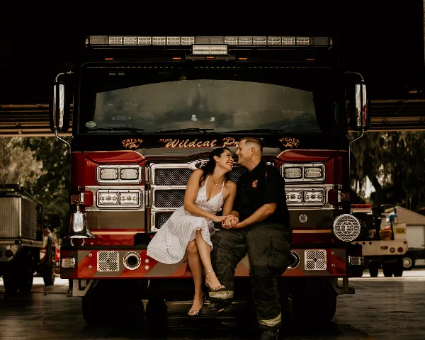 A couple sitting on the bumper of a fire truck, smiling at each other.