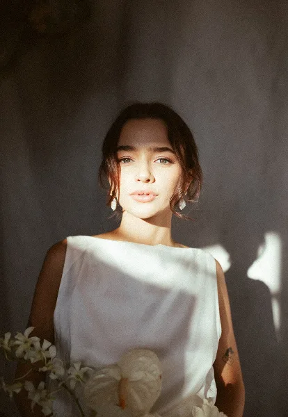 Portrait of a woman in a white sleeveless dress holding white flowers, lit by soft natural light against a dark textured wall.