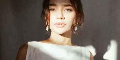 Portrait of a woman in a white sleeveless dress holding white flowers, lit by soft natural light against a dark textured wall.