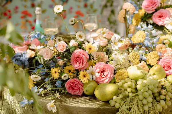 A lush wedding floral arrangement featuring pink roses, white daisies, and green grapes on a gold table.