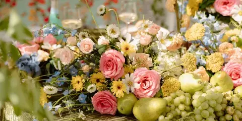 A lush wedding floral arrangement featuring pink roses, white daisies, and green grapes on a gold table.