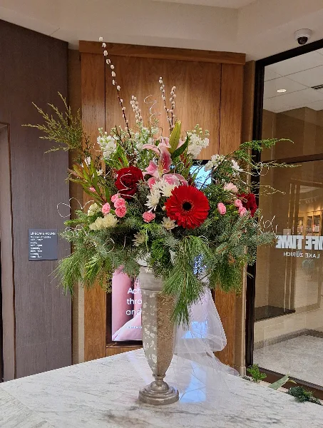 A tall, elegant floral arrangement featuring red gerbera daisies, pink lilies, and greenery in a silver vase.